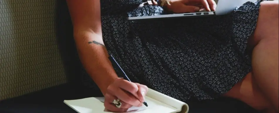 A person typing on a laptop and writing in a notebook.