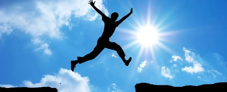 A photograph of someone jumping, with the sun behind them in the blue sky.