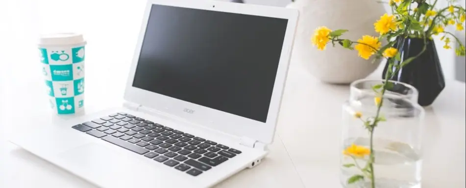 A laptop with a coffee cup and a flower in a vase next to it.