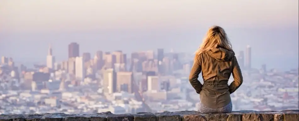 A woman sits on a wall looking out at a city.