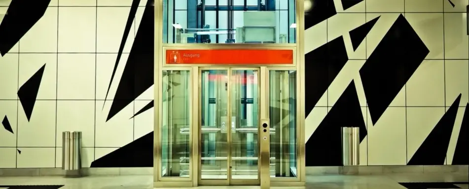 Photograph of an elevator in a big building. There a red strip of paint at the top of the elevator, which is glass, and a black and white design on the wall in the background.