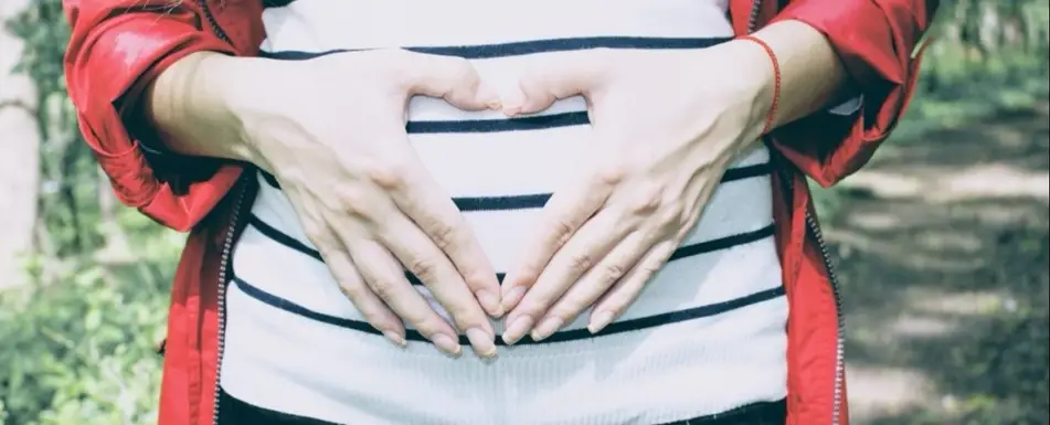 A woman forms a heart with her hands over her pregnant stomach.