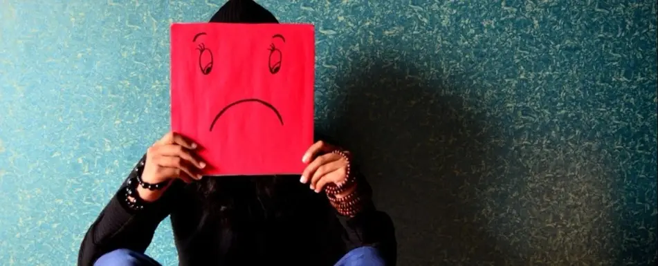 A person holding up a sign with a frowny face, in front of their face.