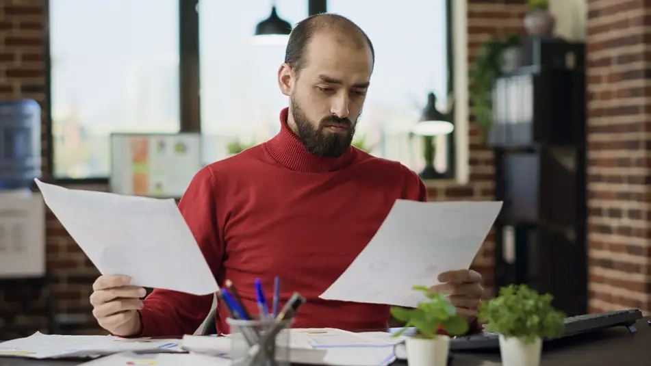 A man wearing a bright red turtleneck shirt sits at his desk reviewing his resume in an office environment.