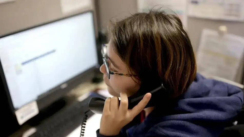 A young person sitting at the computer with a phone to their ear.