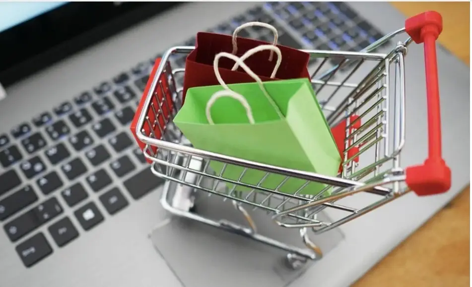Imagem um mini carrinho de compra com duas sacolinhas feitas de dobradura de papel, uma verde limão e outra vermelha escura, em cima de um teclado de um notebook.