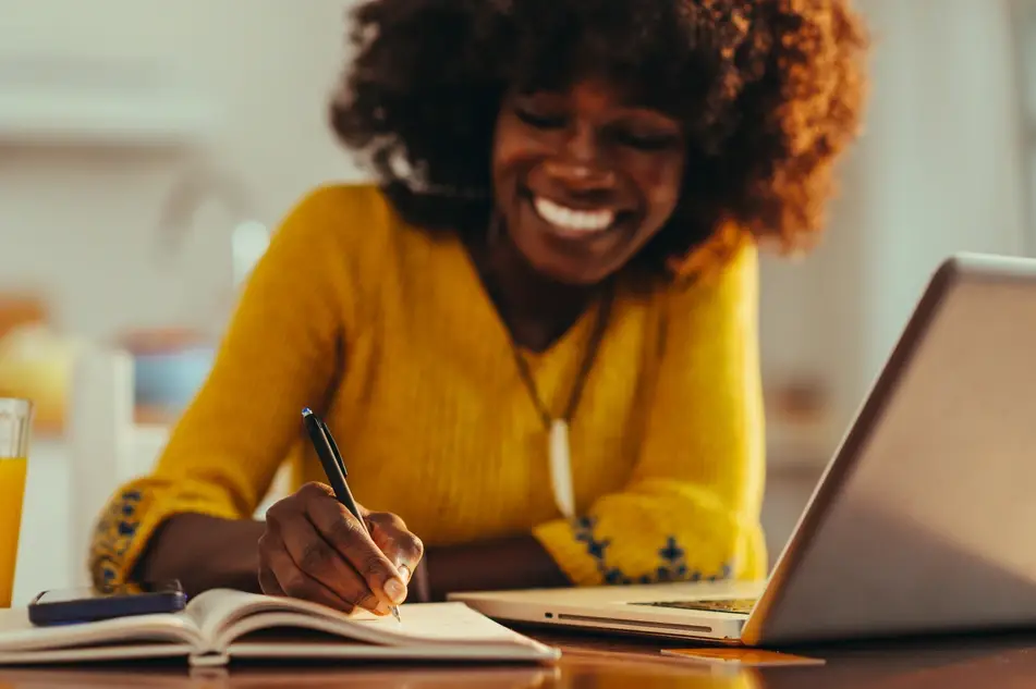 An image of a Black woman in a yellow sweater smiling as she writes in a notebook with a ballpoint pen. Her silver laptop is open on the table next to her, and there's a glass of orange juice next to her right arm.