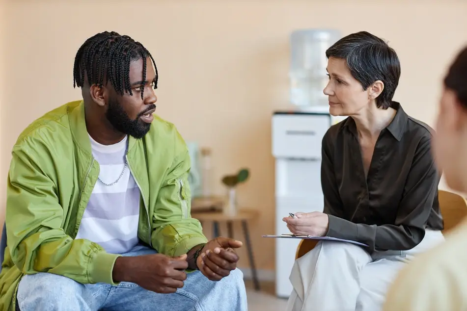 A photograph of a Black man sitting in a chair and speaking to a group of people during a group therapy session, with a water cooler in the background.