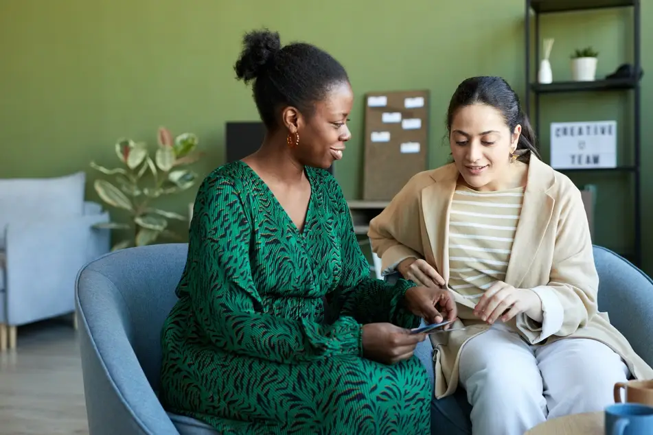 A Black woman offers mentorship to another woman as they sit in an office room with green walls and talk.