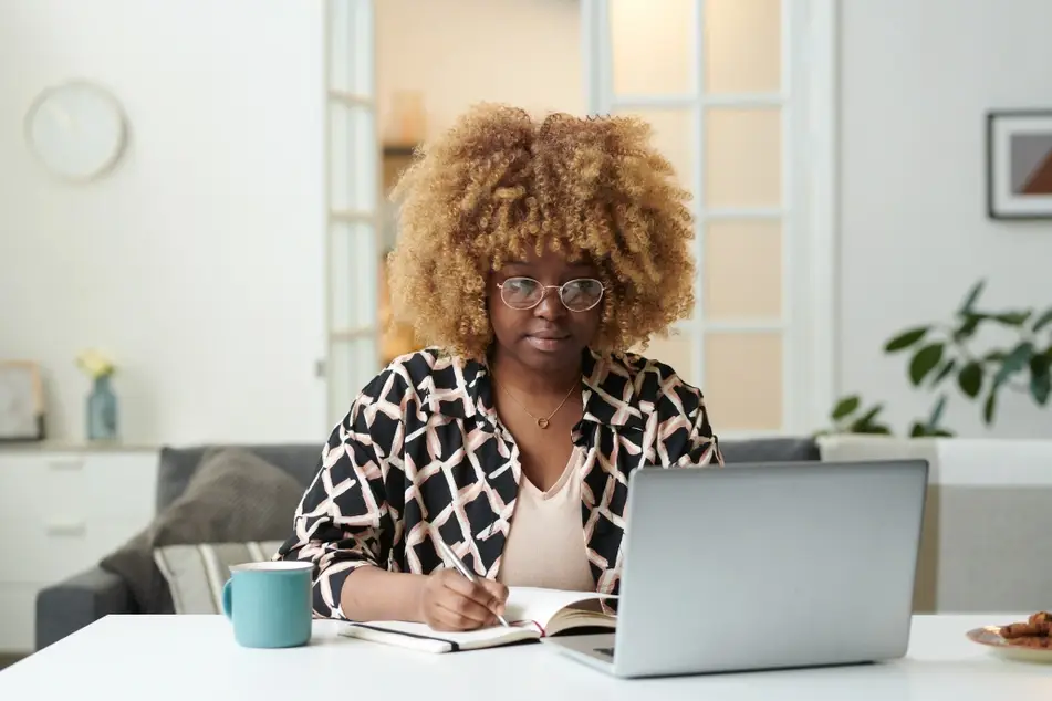 A Black woman sits at a desk in the middle of her living room. Her laptop is open in front of her and she is writing in a notebook, related to the Idealist blog post "The Art of Designing Your Career."