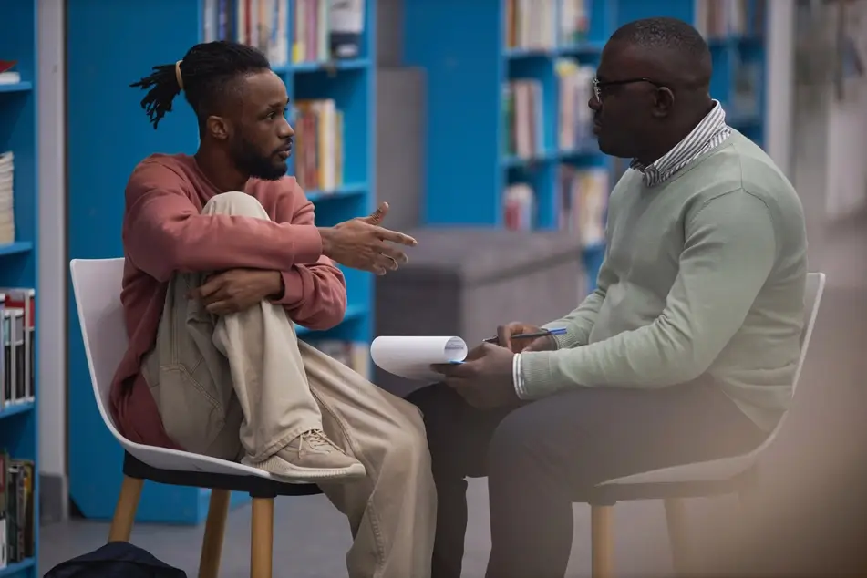 A Black man in a light green sweater sits across from a younger Black student in a library and writes on a clipboard.