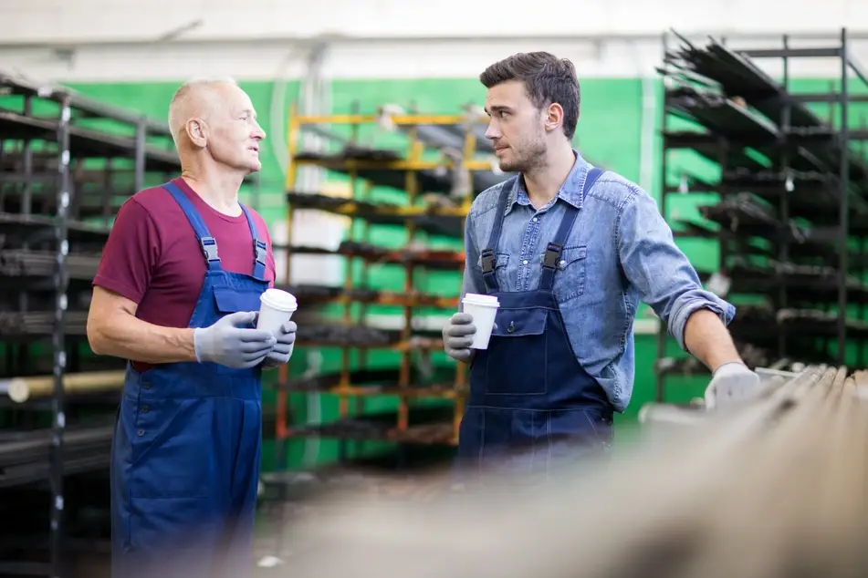 Two white men who work at a warehouse are talking. They're both wearing protective gloves and aprons while holding small white coffee cups. In the background there are rows of shelves holding long metal rods.