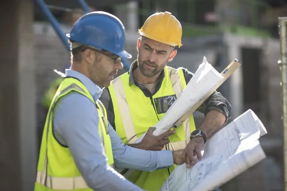 Two white men working at a construction site wear yellow vests, as well as a yellow and blue hat, respectively, as they look over project plans together. One of the men looks at the other inquisitively as he manages up.