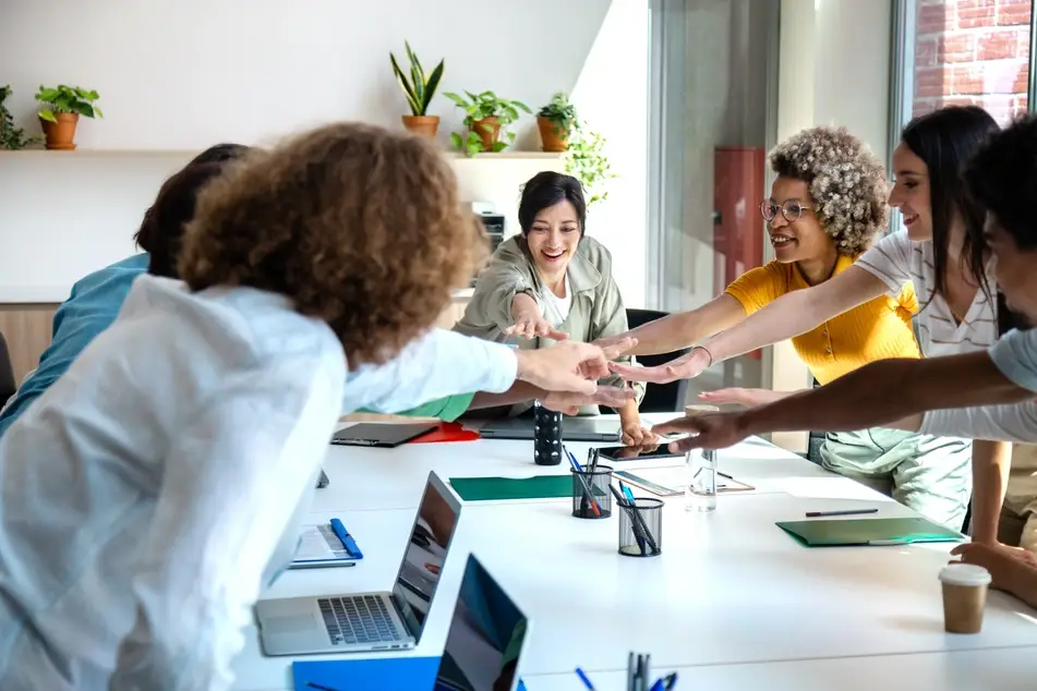 A group of multiracial co-workers are sitting around a long office desks and putting their hands in the middle of the table to signify, "go team!"