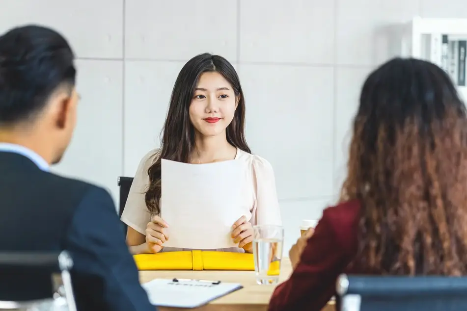 A young woman of Asian descent sits at a long table in front of two hiring managers—a man with short hair and a woman with long curly hair—holding a piece of paper in front of her.