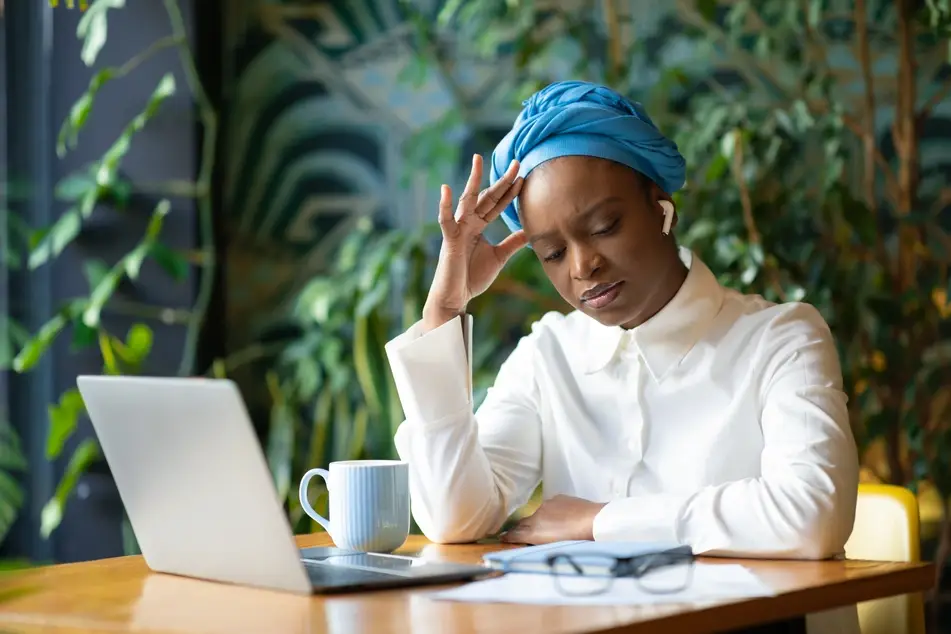 A Black woman appears stressed while sitting at a desk with a laptop, mug, and various papers strewn around her.