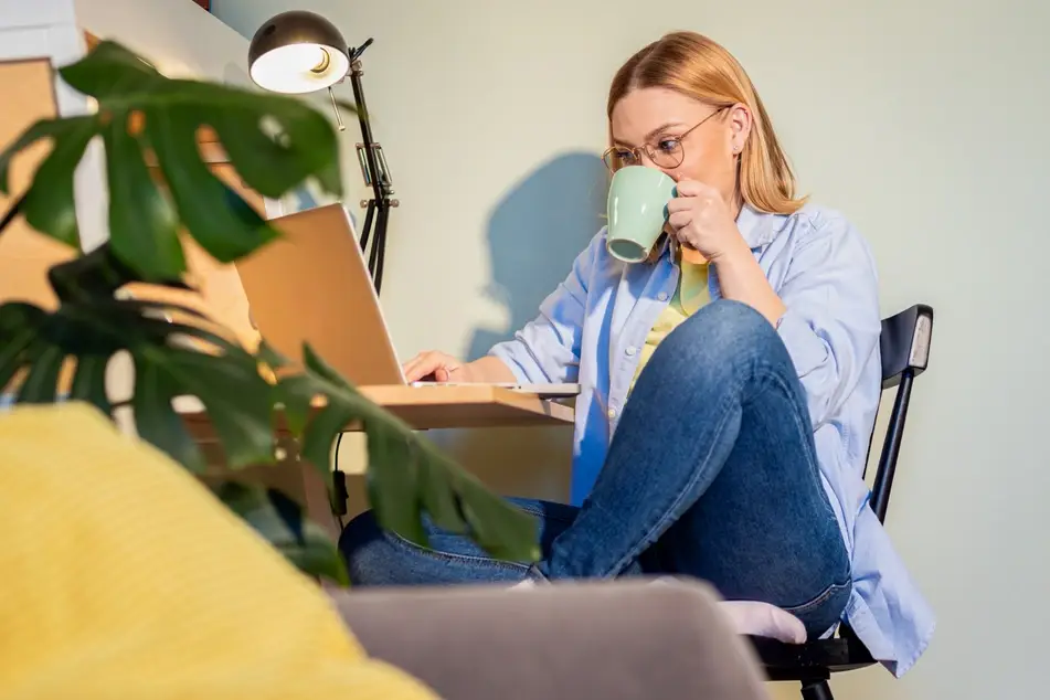 A photograph of a white woman sitting at her desk at home and drinking coffee as she looks at her laptop.