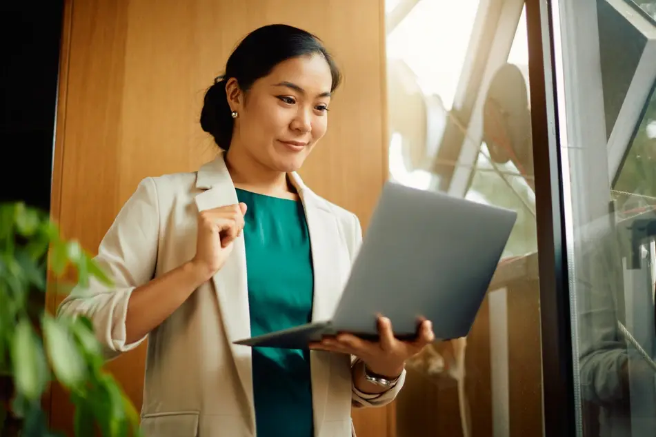 A woman of Asian descent peruses online professional development opportunities on her laptop while standing in her office.
