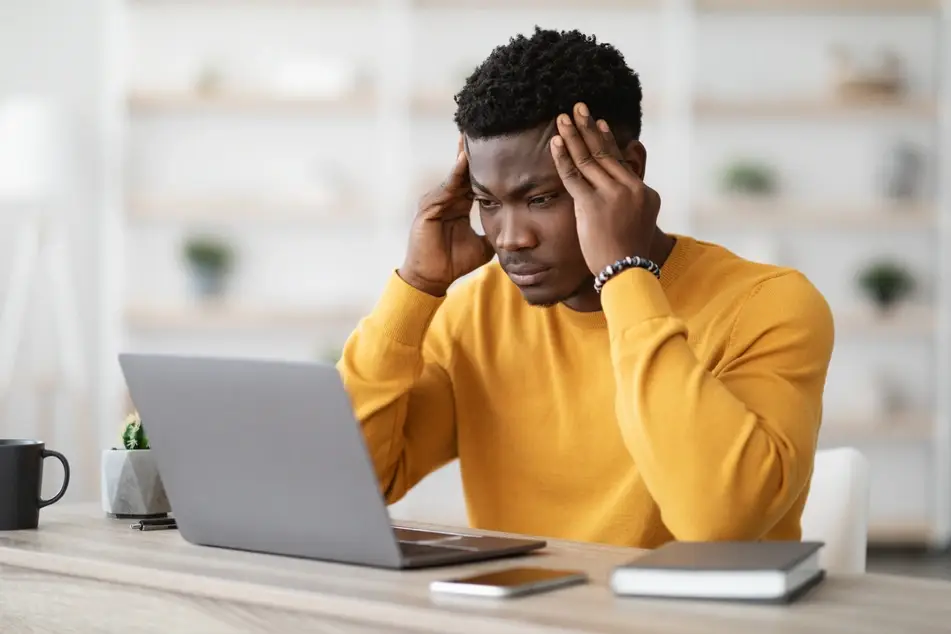 A photograph of a Black man in a yellow sweater startng at his laptop.