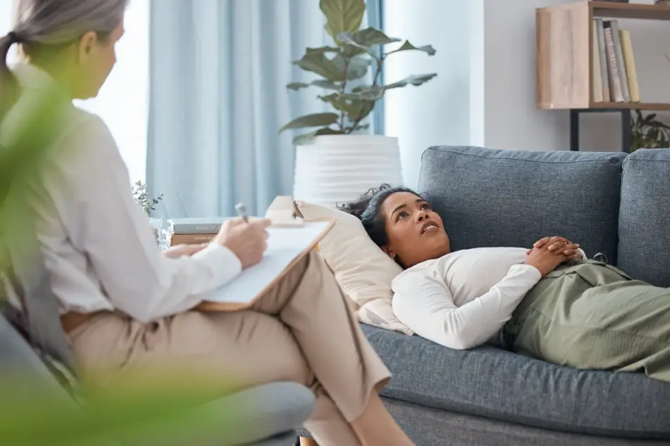 A photograph of a Black woman lying on a couch as she talks to a therapist.