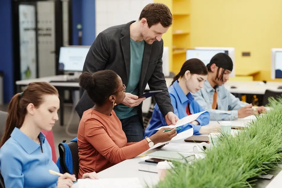 A photograph of several people sitting at a long desk in a colorful university classroom. The teacher, a white man, is reading the paper of one of his students, an older Black woman, while other students continue working.