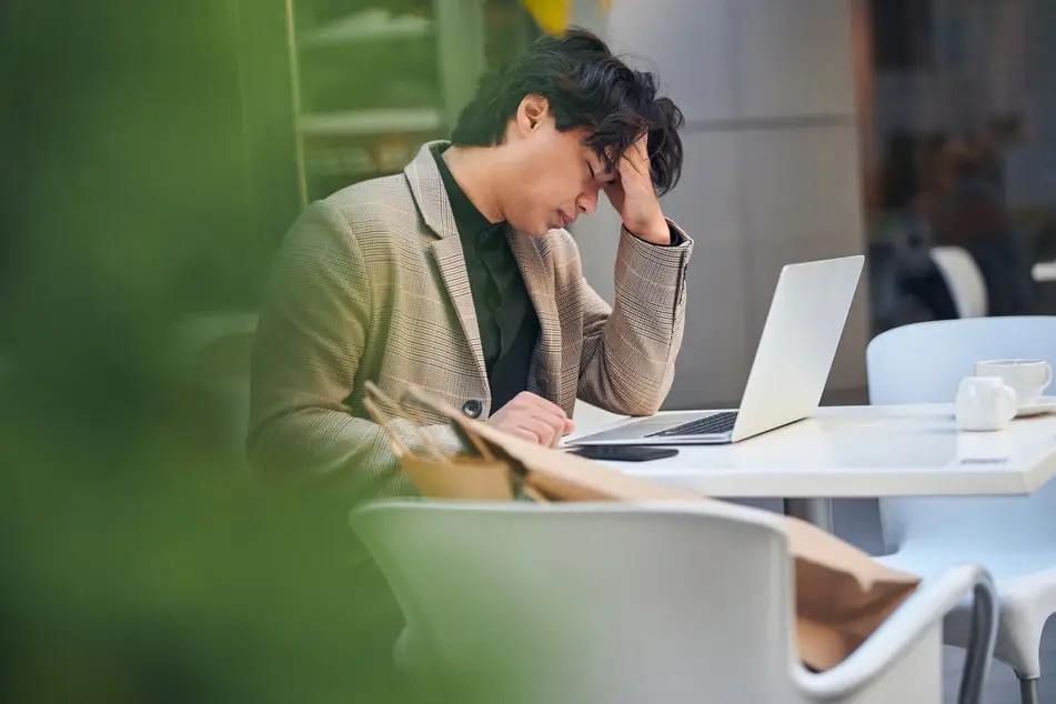 A man with shaggy dark brown hair sits at a table and holds his head in his had, signifying that he is depressed at work. His laptop is open in front of him and there is a green plant blocking the left side of this image.