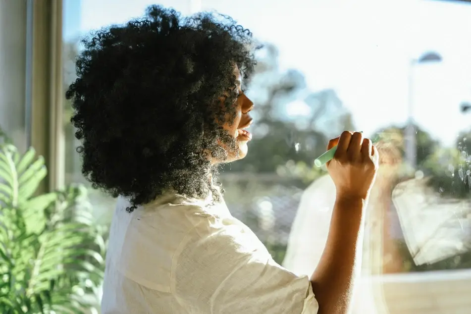 A Black woman wearing a white shirt writes New Year's Resolutions on a window using a green dry-erase marker. There's a green plant in the background.