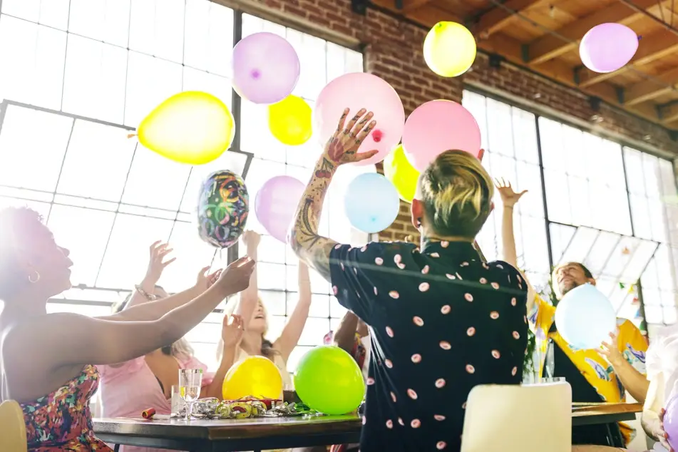 a group of co-workers sit at a long office table having fun at work with colorful baloons.