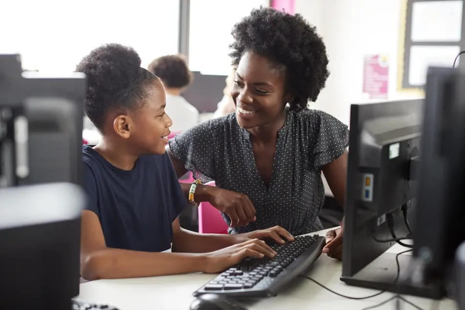 A Black mother and daughter smile at each other while sitting by a computer.