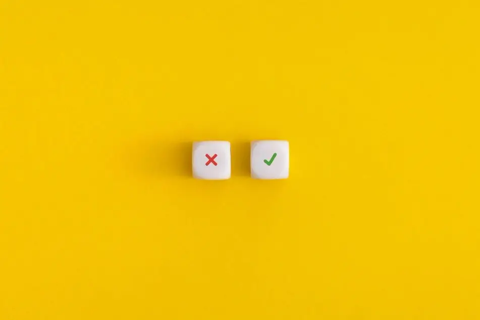 Uma fotografia de dois dados - um com um "x" vermelho e outro com uma marca de seleção verde - em um fundo amarelo, representando o que é certo ou errado nas buscas de emprego.