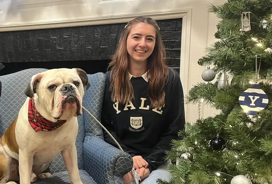 A photograph of Oana Groza wearing a blue Yale University sweatshirt and posing with the bulldog Dan, Yale University's school mascot, next to a Christmas tree.