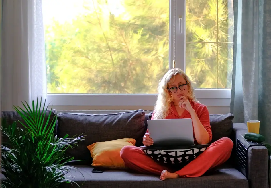 A photograph of a white woman with blonde hair sitting on her couch and working on a laptop.