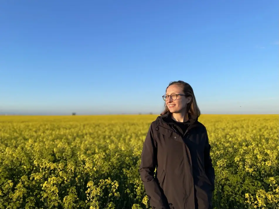 A photograph of Alex Malikova, standing in a field of yellow flowers in Sweden.