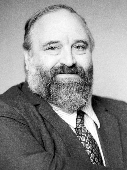 Black and white photo of man with suit and black and gray beard looking at camera.
