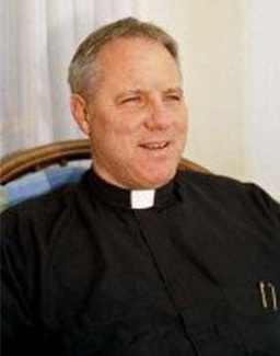 Short haired priest sitting in a blue chair in black outfit with white collar