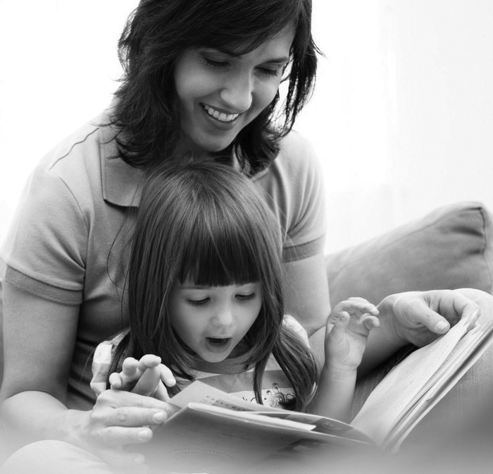 Black and white image of smiling mother reading a book to young girl sitting in her lap