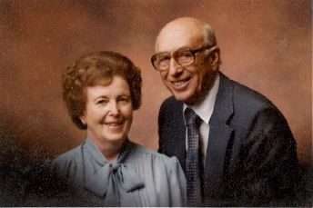Vignette of woman in blue blouse with large bow and man with suit and large black glasses smile at the camera