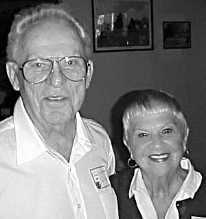 A tall man with glasses and white shirt and a shorter blonde woman smile at camera