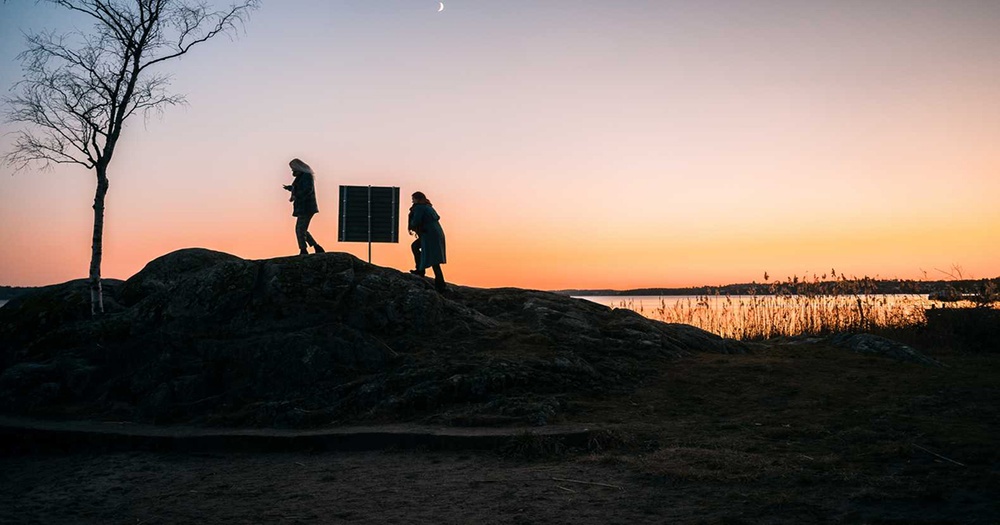 Landskap i skugga där två personer går förbi en skylt.