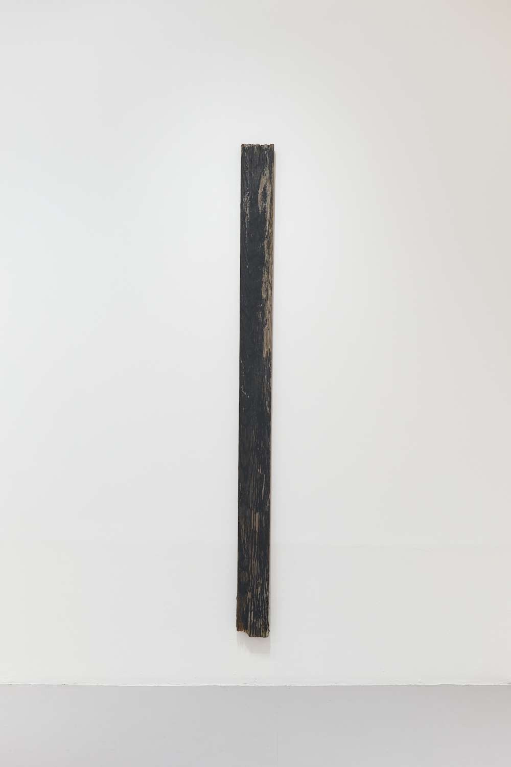 A long, thin, vertical artwork. It is of a plank of wood painted black with original wood grain peeking through and rough cuts on the top and bottom.