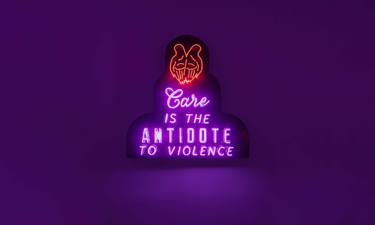 Red neon of two hands palms facing out with purple neon below reading "Carie is the antidote to violence"