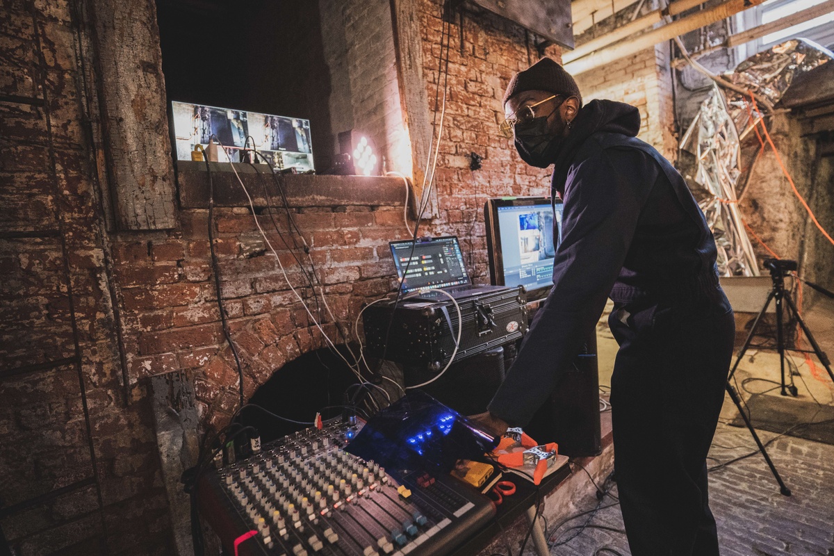 A Black person leans over a sound board and computer surrounded by monitors in a brick basement.