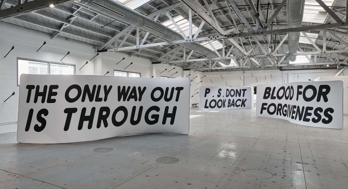 Three-dimensional, white soft sculptures in the shape of a rectangle with a gentle s-curve, staggered throughout the space with black text reading, "the only way out is through", "blood for forgiveness", and "P.S. Don't look back".