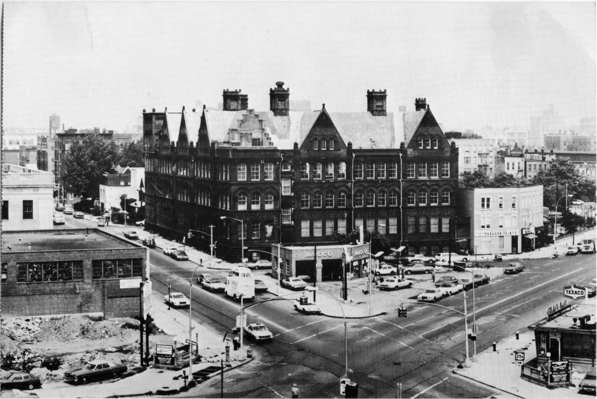 A black and white photo of the brick MoMA PS1 building from a frontfacing aerial view. The building is framed by an intersection of cars, gas stations, and shorter buildings.