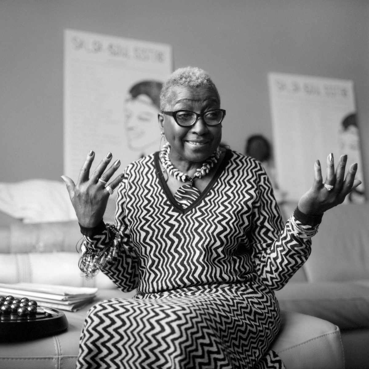 This is a portrait of Cassandra Grant. She's an older Black woman with short hair and glasses. She's wearing a long-sleeve patterned dress and smiling. 