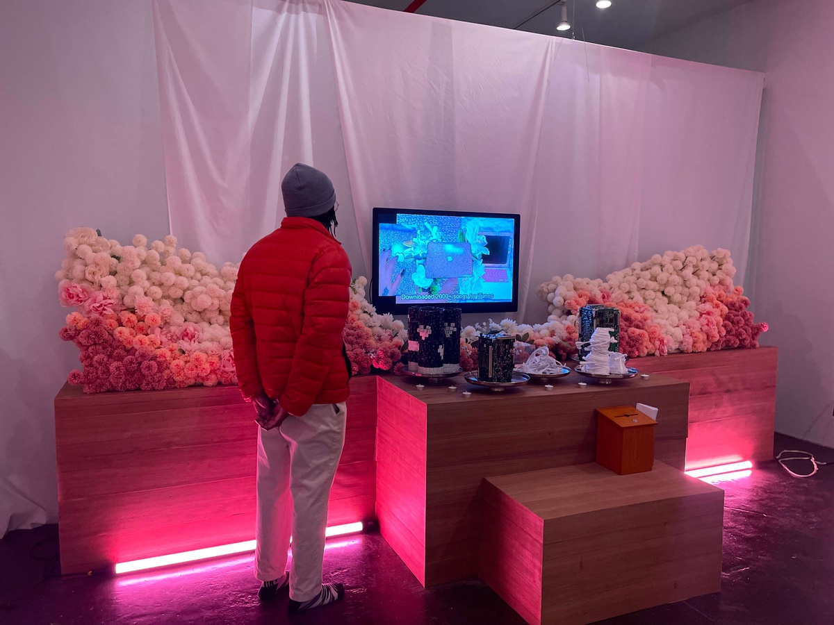 Person stands in front of an altar with their hands behind their back. Atop the altar sits a monitor, pieces of keyboards and circuit boards, and two large bunches of flowers. Beneath the altar glows pink florescent lighting.