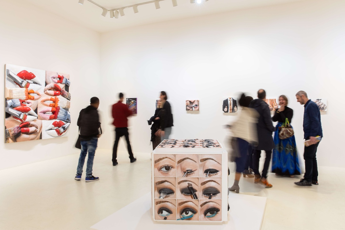 A white-walled gallery space with moving people viewing artwork. A cubed sculpture occupies the foreground, which displays a grid of eyes applying eyeliner and eyeshadow. 