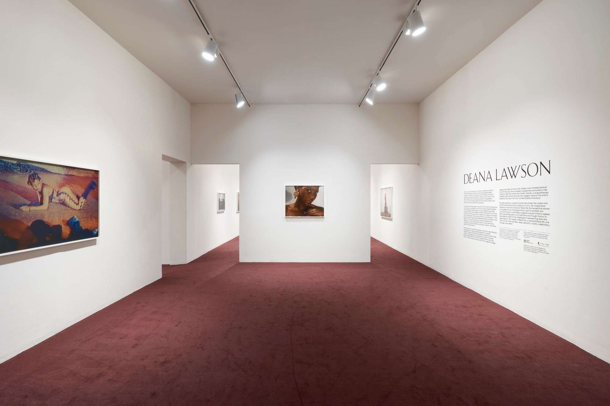 A white wall gallery space with wine colored carpeting. On one wall is a photograph of a Black woman wearing makeup. To her right, the exhibition wall text.
