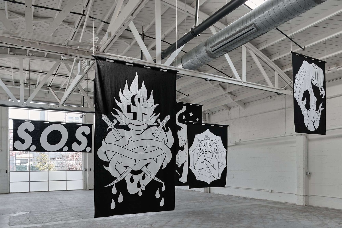 Oversize black banners with white appliqué patterns of a sacred heart, a figure with a spiderweb, a hand with a chair and spiked ball, and text banner spelling out S.O.S. Banners are suspended in the air in a large white warehouse with light gray floors