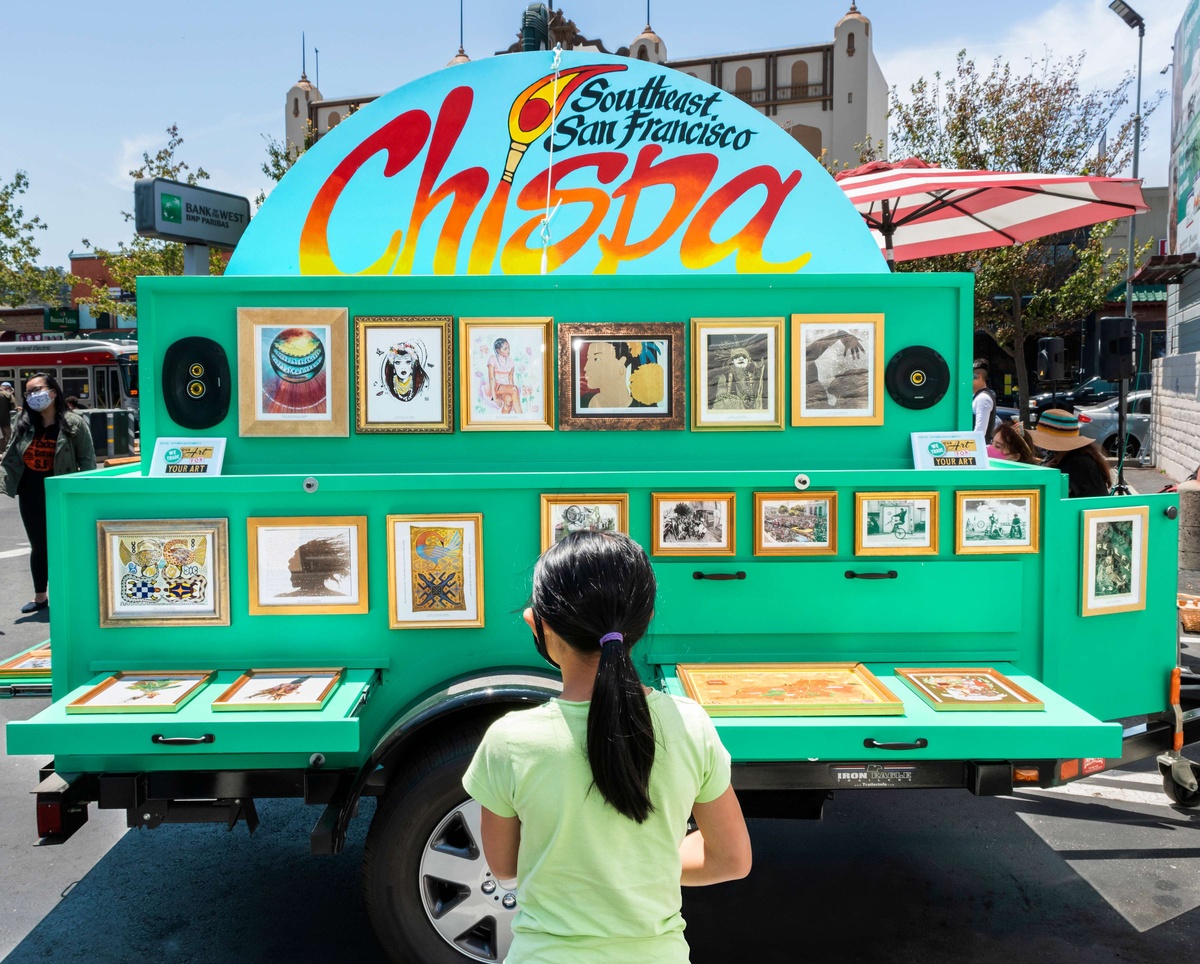 Child in a brigh green shirt with a ponytail facing a large towable art cart covered in art
